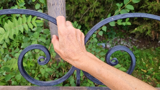 An elderly woman holds on to a metal decorative element on a wooden bench in a park