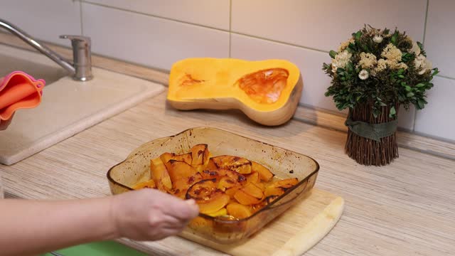 Woman hands in oven mitts add honey and butter from spoon to baked, cooked butternut squash slices in glass container