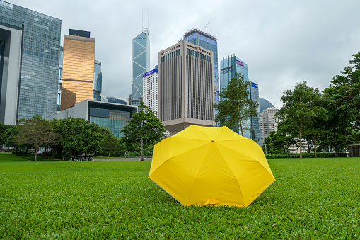A yellow umbrella placed in Tamar Park, Admiralty, Hong Kong Island.  In the background are skyscrapers including the Chinese People's Liberation Army Hong Kong Garrison Building, identified by the star at the top of the building; the Bank of China Tower; and Central Government Complex of the Hong Kong Special Administration Region.  A yellow umbrella is a symbol that arose from democracy protests in 2014 and 2019.  This image was taken on an overcast afternoon on 14 September 2023.