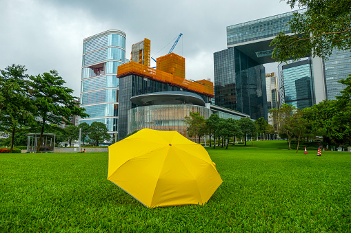 A yellow umbrella placed in Tamar Park, Admiralty, Hong Kong Island.  In the background are buildings the Central Government Complex of the Hong Kong Special Administration Region, including the Legislative Council Complex directly behind the umbrella.  A yellow umbrella is a symbol that arose from democracy protests in 2014 and 2019.  This image was taken on an overcast afternoon on 14 September 2023