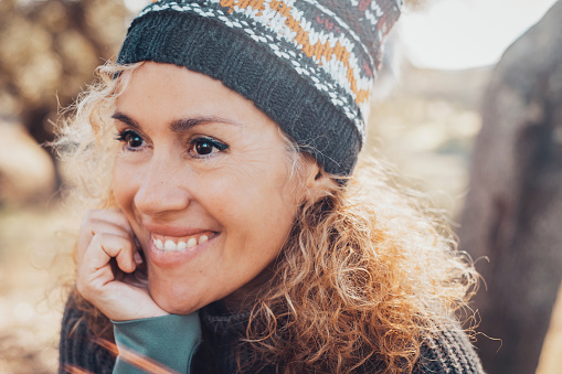 Portrait of cute happy and serene woman middle age smiling and looking in front of her enjoying time and outdoor leisure activity at the park during autumn season. Wearing knit hat. Perfect teeth