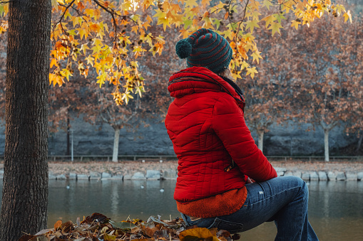Back view of woman with red jacket admiring calm water lake river in autumn season. People having relax outdoor leisure activity alone. Wellbeing and natural lifestyle. Yellow leaves and nature around
