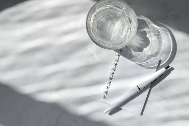 Water glass, blank notebook mockup, pen on white marble table background with natural dappled light shadow. Minimalist aesthetic neutral monochrome business branding template.