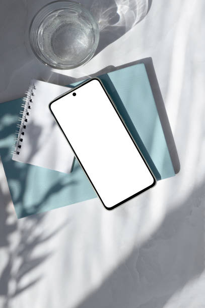 istock Smartphone empty screen mock up, blue notebook, glass with water on white table with natural sunlight shadows. Aesthetic business brand template, copy space 1681896420
