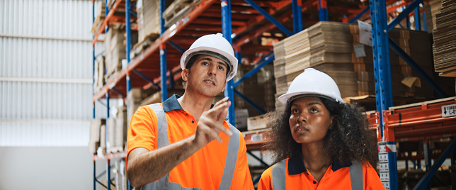 Young adult African American woman, Caucasian man engineer worker work together in factory warehouse, talk explain job process discussion. Logistic industry business, people at industrial work concept