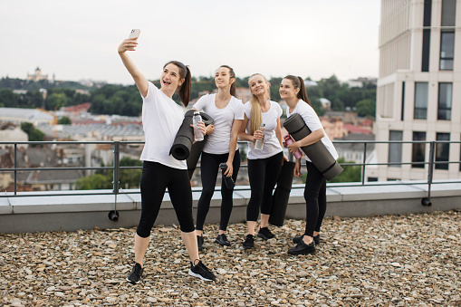 Four excited females with mats and sports bottles taking selfie on mobile phone while relaxing after outdoor workout. Delighted yoga practitioners saving merry moments of time spending together.
