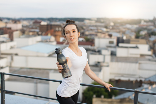 Close up view of pretty lady in activewear reaching out sports bottle while holding handrails on building roof. Young fit adult renewing water balance after outdoor class in urban neighbourhood.