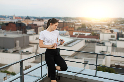 Relaxed health lady in activewear watching city view while leaning on handrails in corner of building roof. Charming yogini with water bottle staring at evening sky after finishing outdoor session.