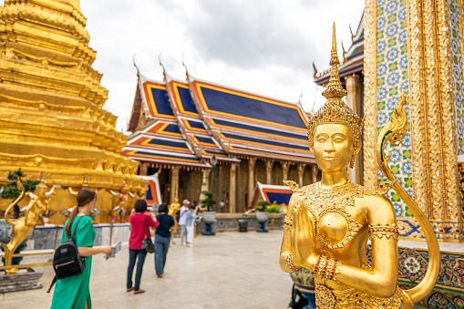 A group of tourists visit the ancient golden temple at Wat Phra Kaew or The Grand Palace, a top tourist attraction in Asia. Bangkok, Thailand. Focus on the golden Kinnari statue.