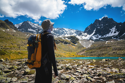 A woman on a mountain with a backpack is watching a beautiful view of the mountains in the background. Hiker girl with backpack in high snowy mountains in summer.