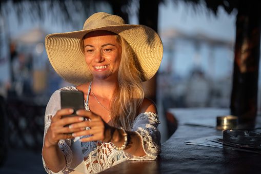 Happy blonde woman whit sun hat holding mobile phone while sitting in cafe' at summer