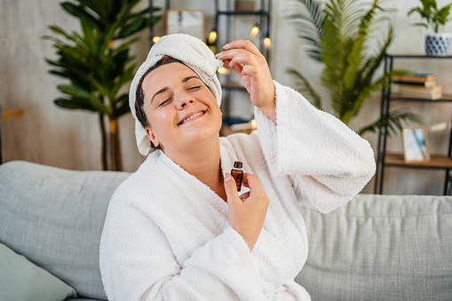 Beautiful young plus size woman in a bathrobe applying face oil in the living room at home.