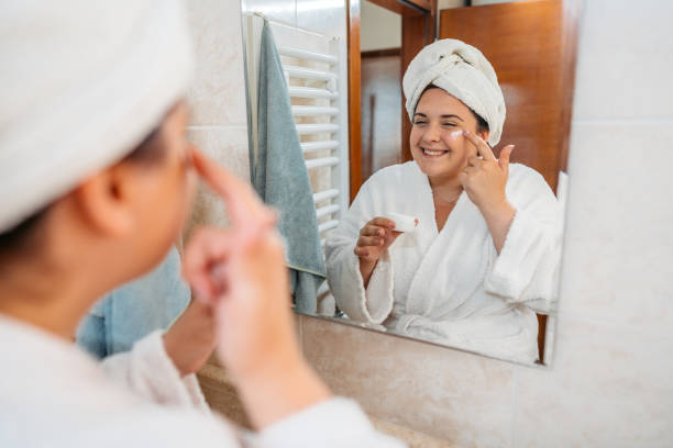 istock Young Woman Applying Moisturizer On Her Face In The Bathroom Mirror 1682065801