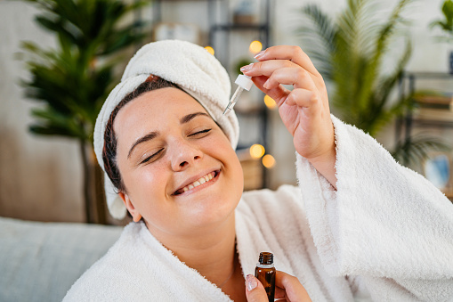 Beautiful young plus size woman in a bathrobe applying face oil in the living room at home.