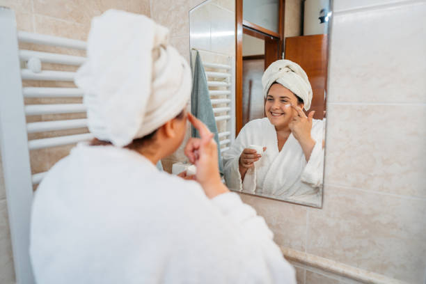 istock Young Woman Applying Moisturizer On Her Face In The Bathroom Mirror 1682073377