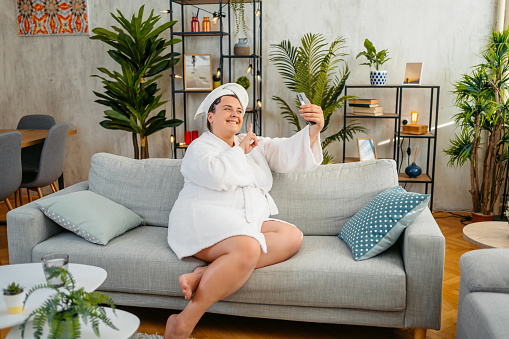 Beautiful young plus size woman in a bathrobe sitting on the sofa in the living room and taking selfies using her smart phone.
