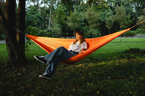 Stunning woman enjoys a peaceful evening, reclining in an orange hammock at the park, relishing a moment of tranquil relaxation amidst nature's beauty