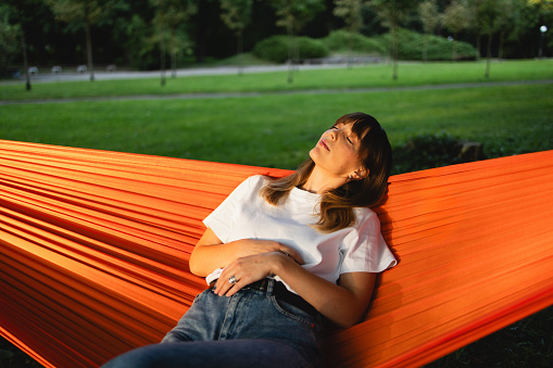 Gorgeous woman unwinds in the evening, her eyes closed in blissful relaxation, swaying gently in an orange hammock at the park, savoring the tranquil beauty of the moment