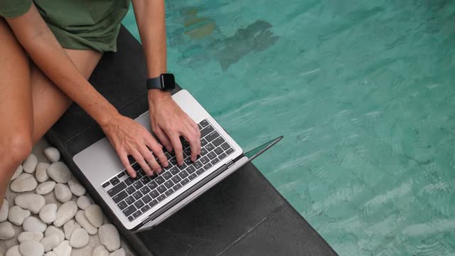 Mid adult woman sitting at the poolside, using laptop