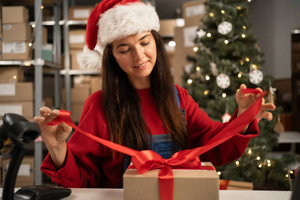 istock Woman packing present box wrapping gift with red ribbon preparing post shipping delivery parcel on table in warehouse. Christmas shipping delivery 1682088512