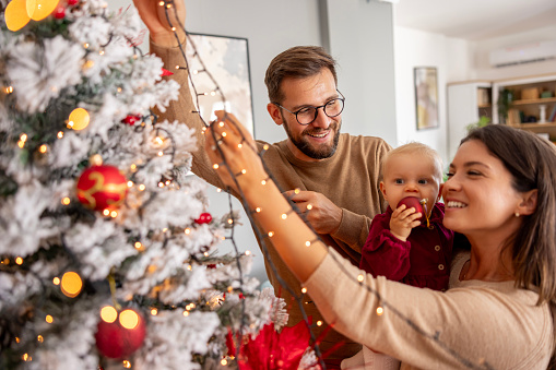 Young parents having fun decorating Christmas tree with their cute little baby girl, placing Christmas lights on it while decorating home for winter holiday season