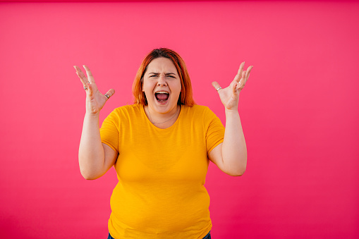 Portrait of an angry young plus size woman standing in front of a pink background and shouting.
