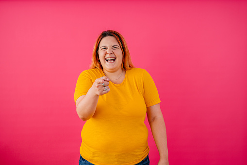 Portrait of a beautiful young plus size woman pointing with her finger at the camera and laughing while standing in front of a bright pink background.