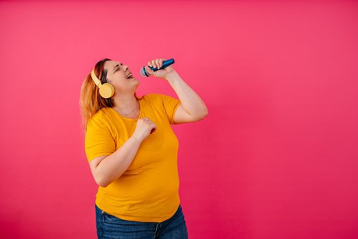 Portrait of a beautiful young plus size woman listening to music via wireless headphones and dancing while standing in front of a bright pink background.