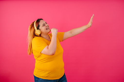 Portrait of a beautiful young plus size woman listening to music via wireless headphones and dancing while standing in front of a bright pink background.