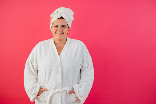 Portrait of a beautiful young plus size woman in a bathrobe standing against a pink background.