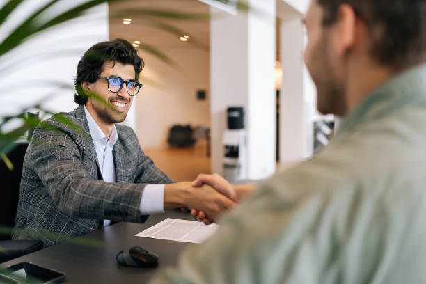 Selective focus of smiling real estate broker in business suit present and advise young man client on decision to sign insurance contract. Selective focus of smiling real estate broker in business suit present and advise young man client on decision to sign insurance contract. Happy customer signing purchase agreement and shaking hands. real estate agent male stock pictures, royalty-free photos & images