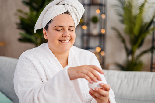 Beautiful young plus size woman in a bathrobe holding a moisturizer while sitting on the sofa in the living room.