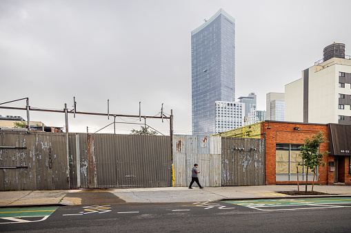 Crescent street, Long Island City, Queens, New York, USA - August 15th 2023:  Man walking in front of a fence in a old small businesses area with a modern residential skyscraper behind