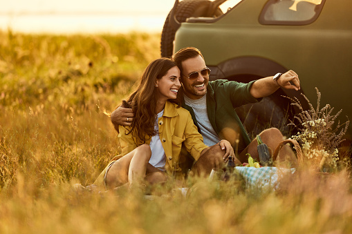 A happy romantic couple having a picnic in nature next to their parked car off-road.