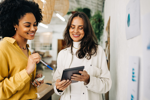 Happy female entrepreneurs using a tablet as they brainstorm ideas for their business. Two business women working as a team to achieve success and profit in their startup.