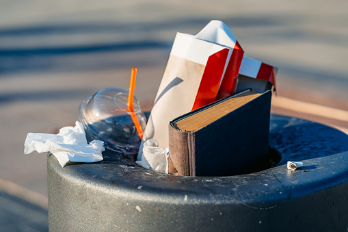A trash can filled with trash (fast food bag, empty disposable plastic cup and a book)