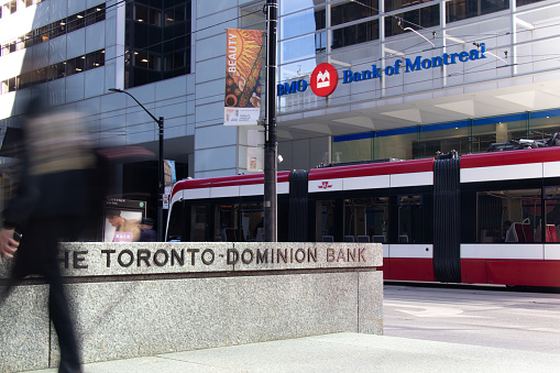 A TTC streetcar passes by the base of BMO's First Canadian Place as a businessman, blurred, walks by a TD Bank sign in Toronto Financial District.