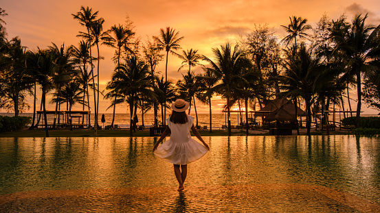 Asian woman watching sunset at a tropical swimming pool with palm trees at the island of Koh Kood Thailand, pool during sunset on the beach at a luxury hotel resort