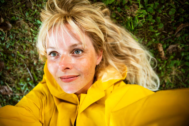 Blonde Woman close up portrait making selfie on meadow. Beautiful Female in yellow raincoat Laying on the grass lawn.