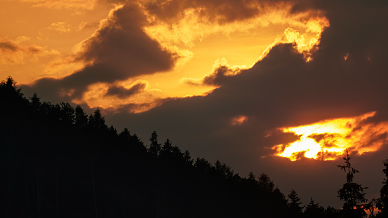 Sun setting down through clouds, above hill with coniferous trees silhouettes