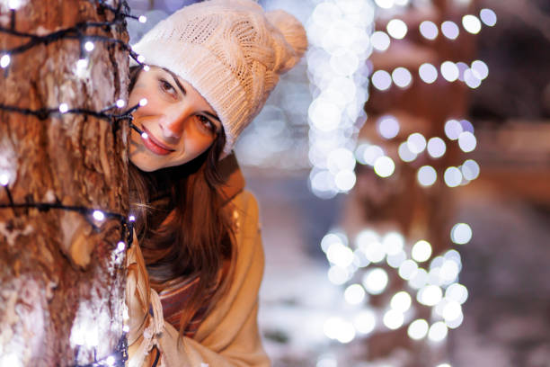 Woman peeking behind tree decorated with Christmas ights while spending Christmas Eve outdoors stock photo