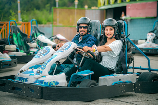 A happy young couple rides a go-cart at an amusement park. They sit together in a cart designed for two people, side by side. They look at the camera with a smile.