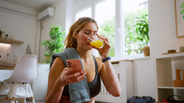 Caucasian woman with vitiligo, using mobile phone and drinking orange juice, after she finished with home workout
