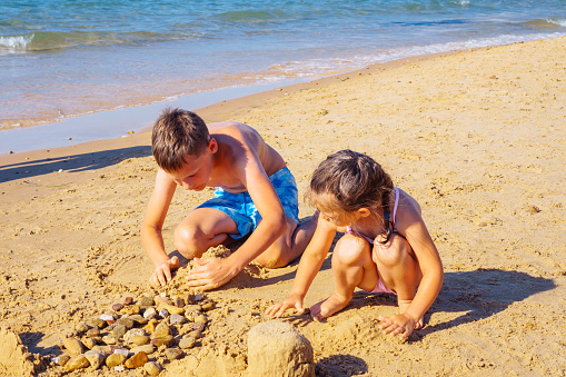 Portrait of preteen boy brother, little girl sister siblings family sitting on beach seashore, playing with sand, building castles near foamy sea waves, stones on sunny day. Summer, travelling, sea.