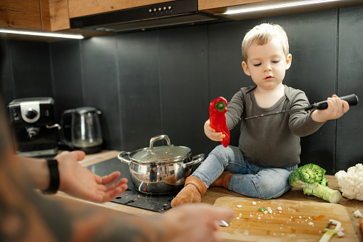 Portrait of amazing little boy toddler sitting on table near cooker in black kitchen at home, holding red pepper and huge knife, father stretching hands to help son. Childhood, cooking, vegetables.