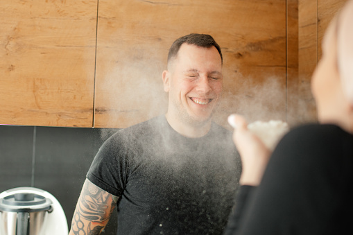 Portrait of joyful young man wear black T-shirt, standing with closed eyes in kitchen at home, fooling with wife. Unrecognizable woman blowing flour at husband face, having fun. Family, relationship.