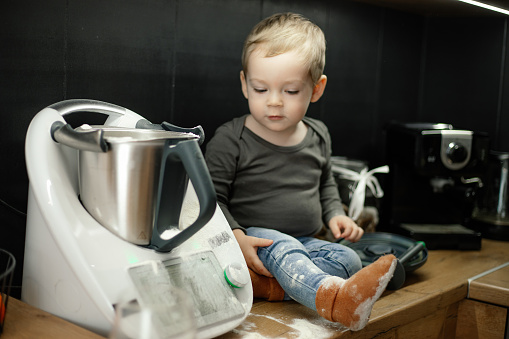 Portrait of cherubic little boy baby toddler with short fair hair sitting on table near kneader cooking machine in black kitchen at home, looking at floury leg. Childhood, cooking, baking, pastry.