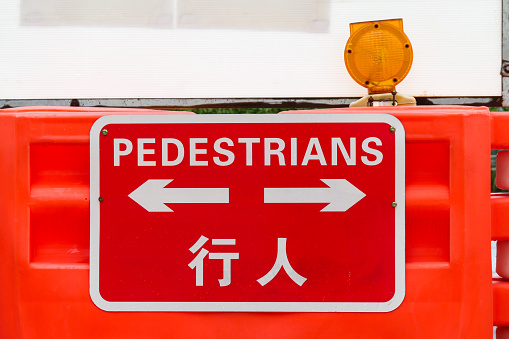 A sign on a crash barrier directing pedestrians in a construction zone outside the Hong Kong Convention Centre in Wan Chai.  Rain droplets are visible on the sign. A white coloured board has been attached to the fence behind the sign. This image was taken on an overcast afternoon on 14 September 2023.