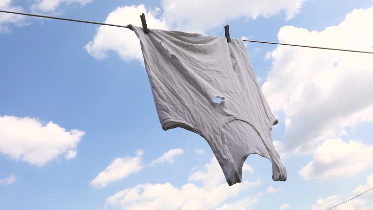 An old white T-shirt with holes hangs on a clothesline against the sky, poverty concept