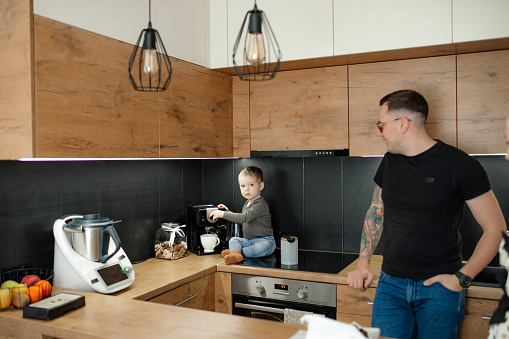 Portrait of family of young man, little boy in kitchen at home. Son sitting near coffee machine, playing with electric kitchen coffee-maker, father leaning on table. Household appliances, technology.